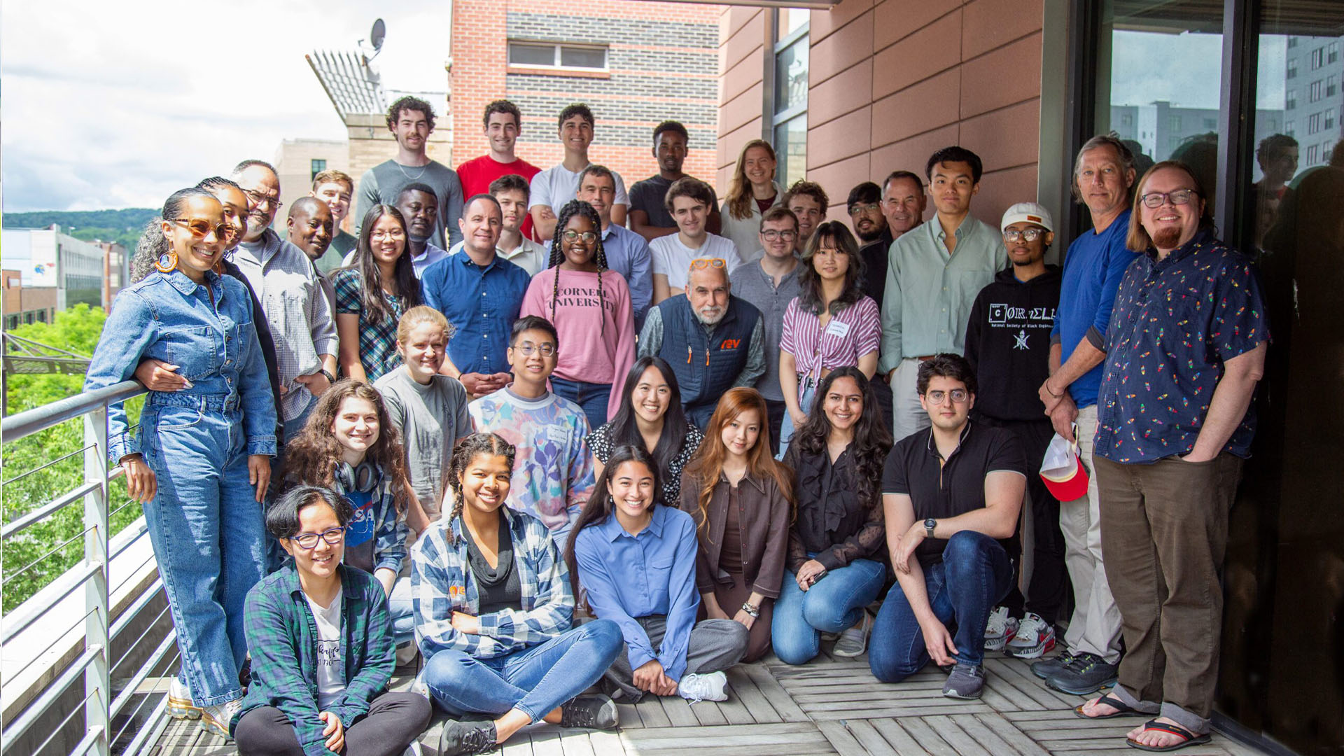 A diverse group of around 30 people is gathered on an outdoor balcony for a group photo. They appear to be a mix of young adults and a few older individuals, all casually dressed. Some are sitting on the wooden deck, while others stand behind them. The background shows a view of buildings and greenery under a cloudy sky. The group is smiling and appears to be enjoying the moment together, with the vibe of a casual, friendly gathering.