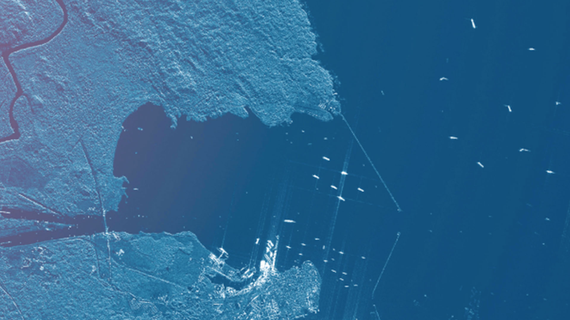 A satellite or aerial image shows a coastal region with a large body of water surrounded by land. The image is rendered in shades of blue, giving it a stylized, almost monochromatic appearance. The land is textured with visible contours, roads, and possibly vegetation, while the water is darker with small white specks that could represent boats or ships. The image seems to highlight the contrast between the land and the water, with a focus on the coastal area and its features.