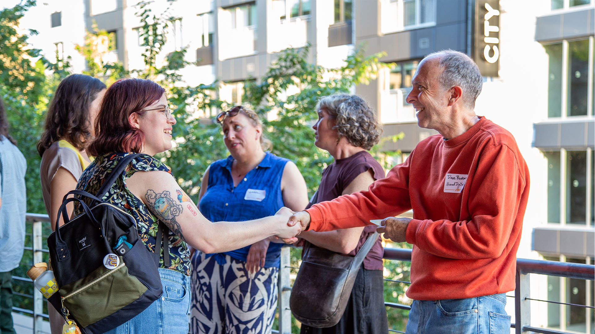 Two people are shaking hands on an outdoor balcony, engaged in a friendly conversation. The person on the left has short, reddish hair and is wearing glasses, a floral-patterned top, and denim overalls. They have tattoos on their arm and are carrying a black and olive-green backpack with various pins and a water bottle attached. The person on the right is an older individual with short gray hair, wearing a red sweater and jeans. Behind them, a few other people are conversing, with a cityscape and greenery visible in the background. The atmosphere appears casual and warm, suggesting a community or social gathering.