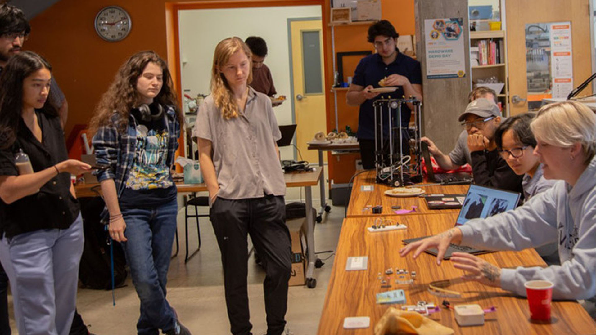 A group of people is gathered in a collaborative workspace, engaged in what appears to be a demonstration or presentation. Several individuals are standing around a table that displays various small objects and devices, possibly related to a technology or hardware project. One person seated at the table is explaining or showcasing something to the others, using hand gestures to emphasize points. The atmosphere is focused, with some participants actively observing and others working in the background. The room is well-lit, with posters and equipment visible, indicating a creative or educational environment, a "demo day".