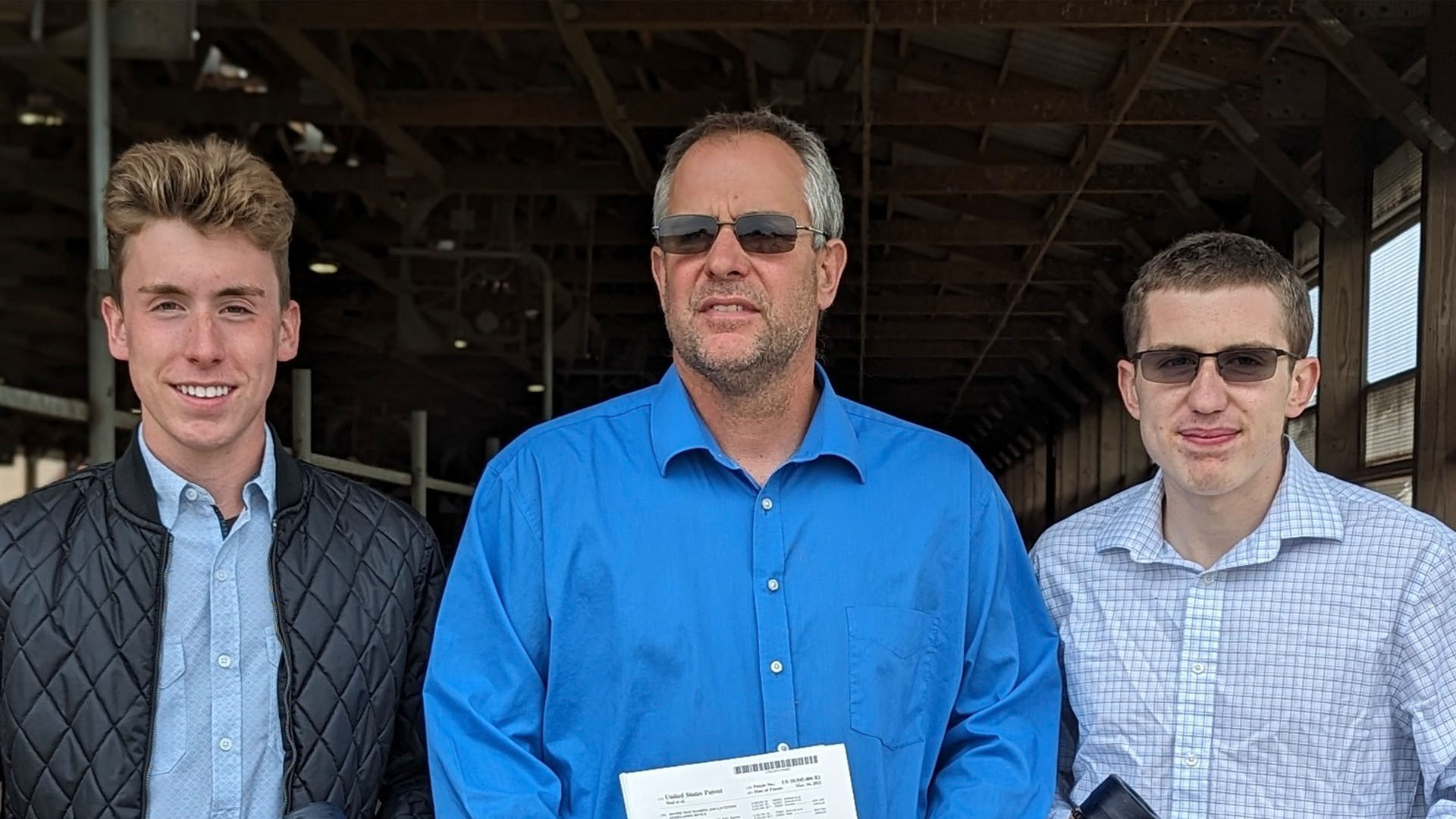 Three men are standing close together in an outdoor setting, possibly under a covered structure like a barn or shed. The man on the left has light brown hair, is wearing a quilted black jacket over a light blue button-down shirt, and is smiling at the camera. The man in the center is older, with gray hair and a beard, wearing sunglasses and a bright blue button-down shirt, holding a document. The man on the right has short light brown hair, is wearing glasses and a light checkered button-down shirt, and has a neutral expression. The group appears to be posing for a photo, perhaps at a family or work-related event.