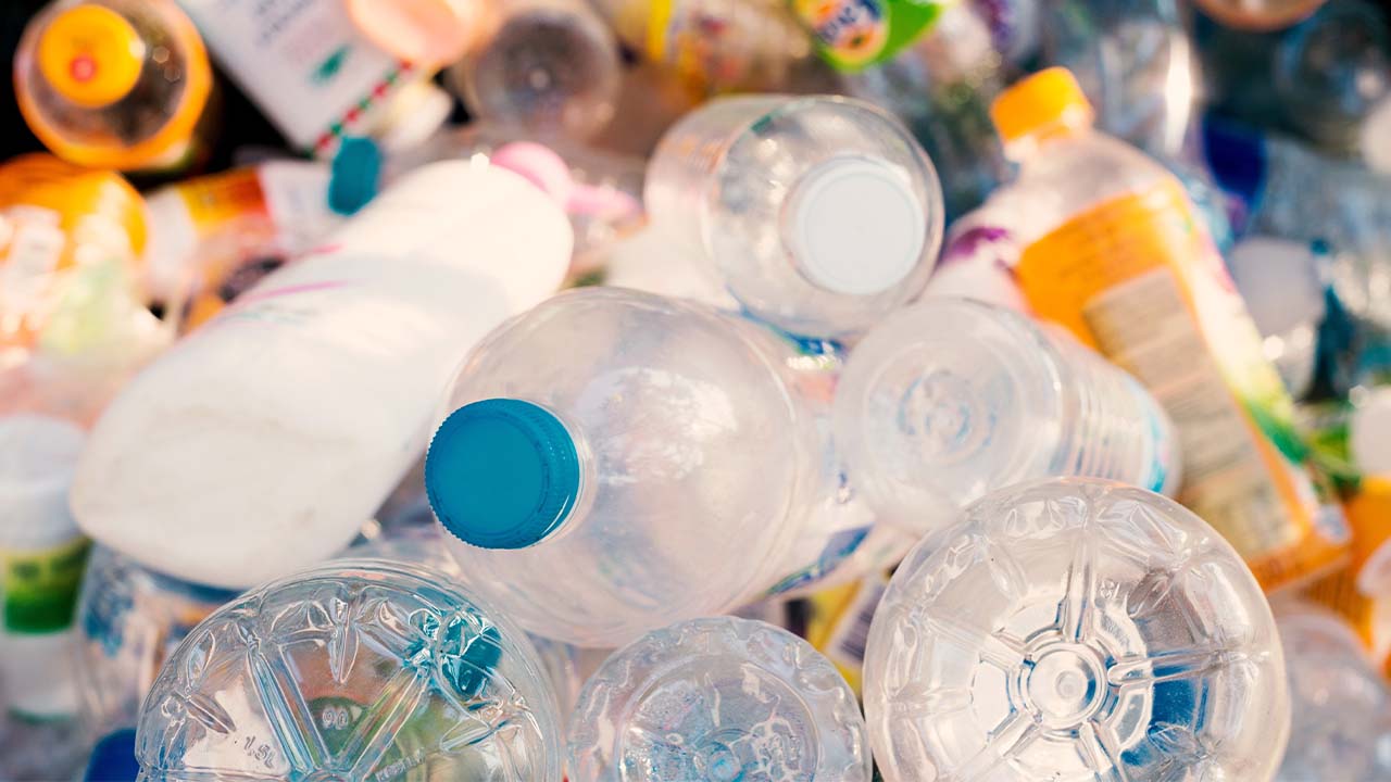 The image shows a large pile of discarded plastic bottles and containers, including various shapes and sizes, some with brightly colored caps and labels. The items appear to be from beverages, with many of the bottles being transparent or translucent. The scene suggests a focus on waste accumulation, particularly related to plastic waste and recycling. The image is brightly lit, highlighting the mixed textures and materials of the plastic items.