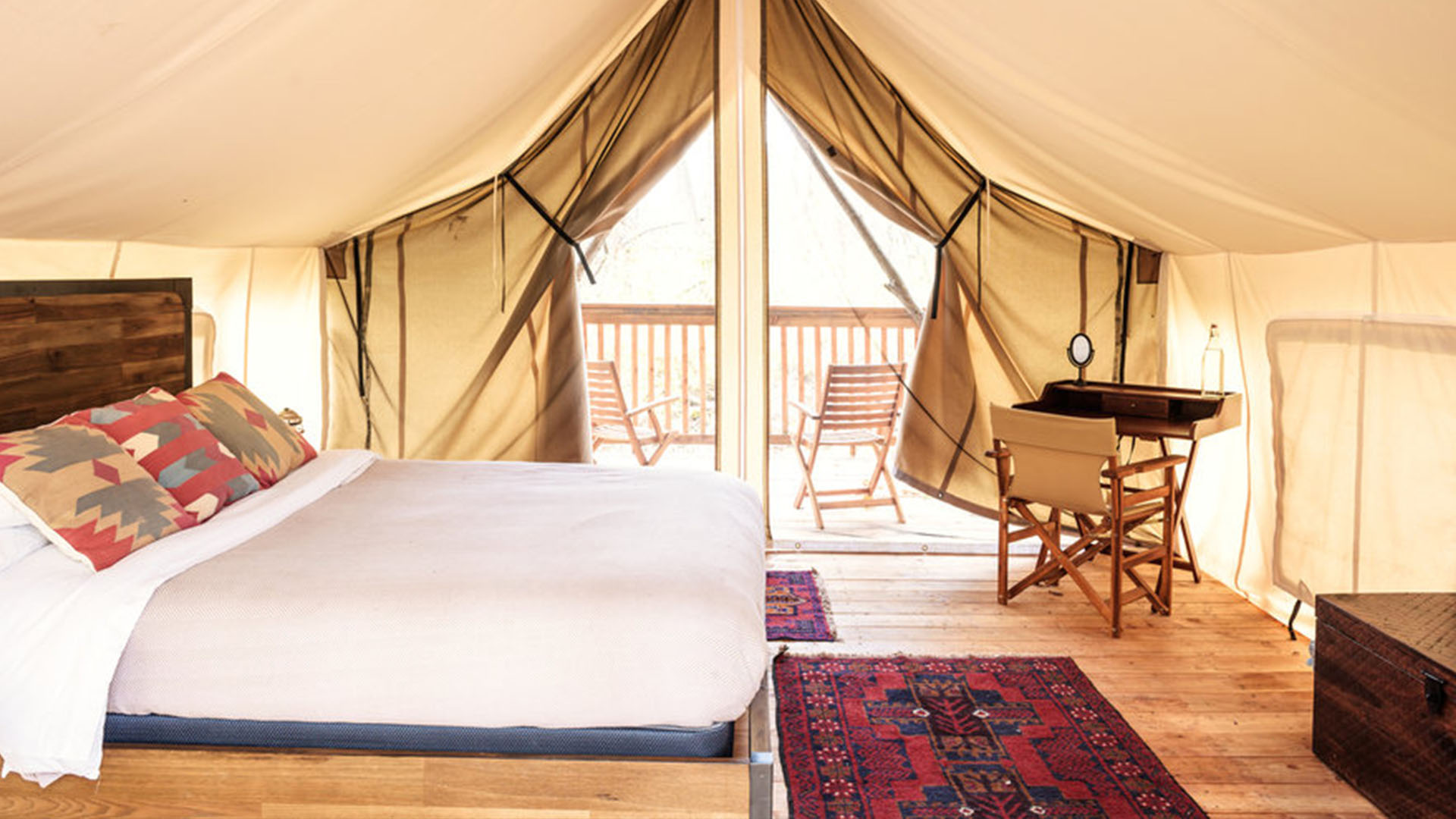 This image shows the interior of a luxurious glamping tent. The tent is equipped with a cozy bed that has geometric-patterned pillows, a small wooden desk with a chair, and a Persian-style rug on the wooden floor. There is a view out to a private deck with two chairs and a table, allowing guests to relax and enjoy nature. The canvas tent is spacious and lets in plenty of natural light, creating a calm and inviting atmosphere.