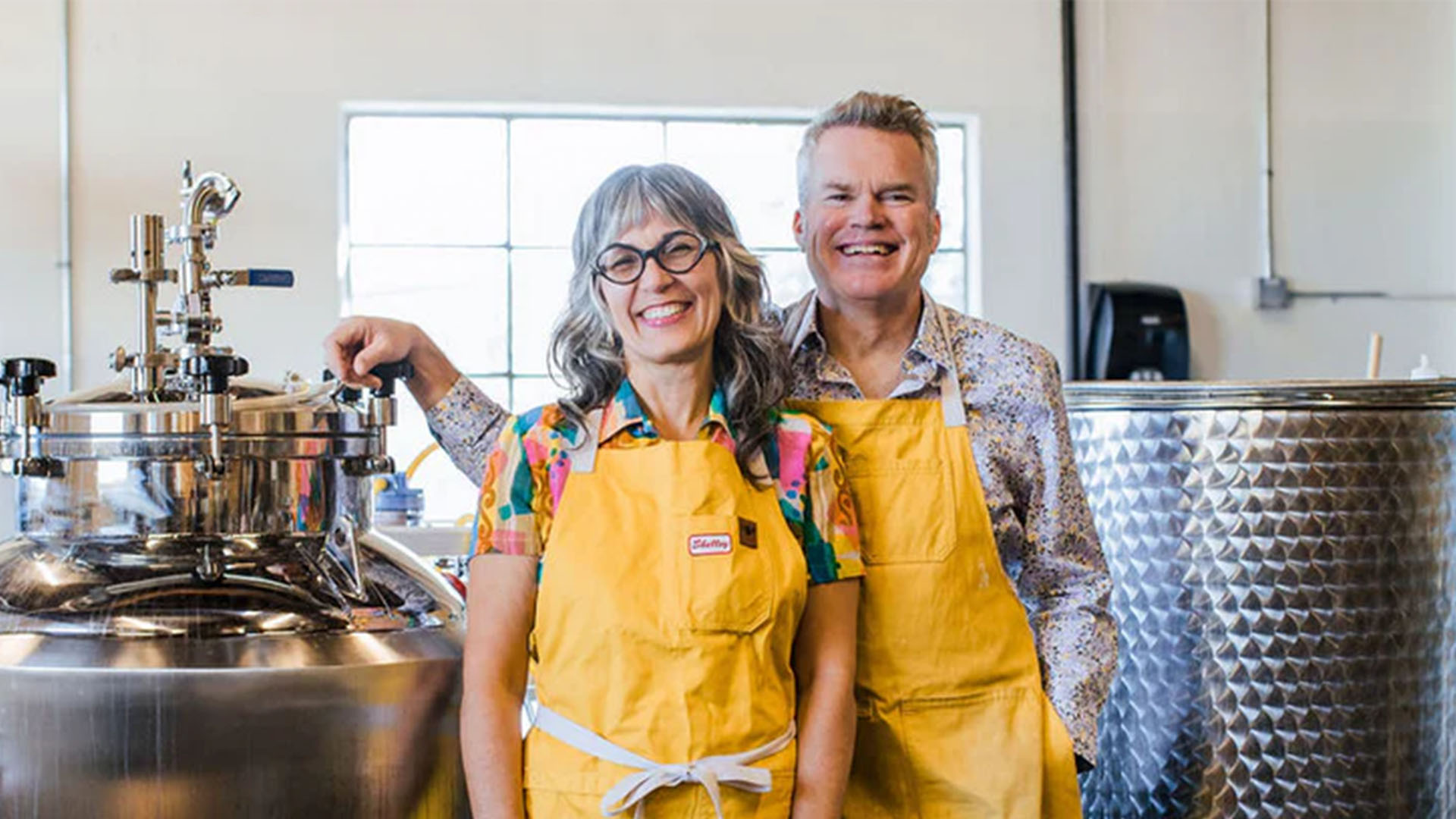 The image shows two cheerful individuals, likely founders or key members of For Bitter For Worse, standing in a modern production setting. They are wearing colorful aprons and are positioned next to industrial equipment, possibly used in the creation of their mocktails. Their warm smiles suggest a sense of pride and enthusiasm for their work.